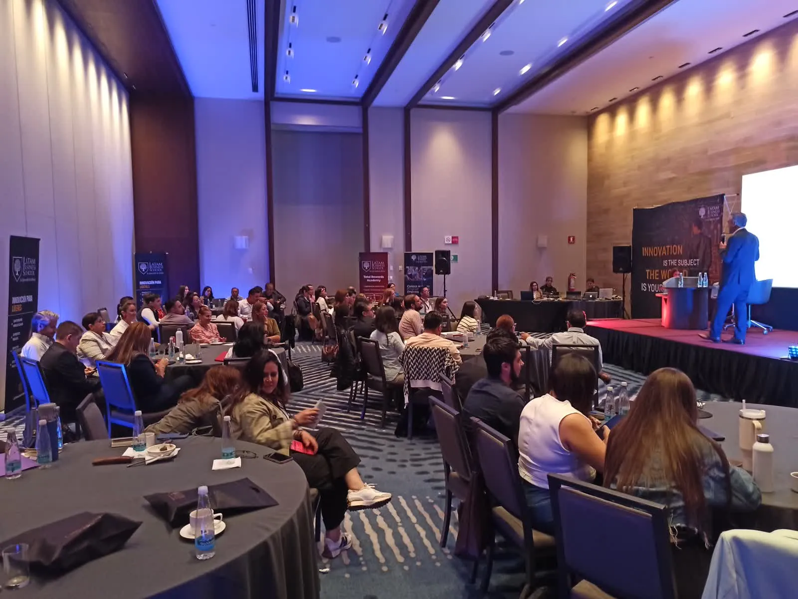 Sala plena de una Cumbre C&B: mesas de trabajo frente al escenario y la pantalla principal
